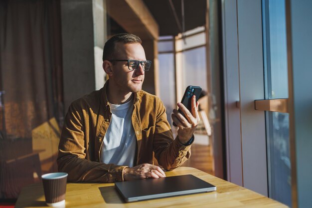 A happy young man wearing glasses and casual clothes is talking on the phone while working on a laptop from a cozy workspace A successful freelancer works remotely