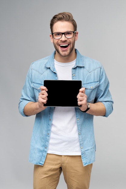 Happy young man wearing jeans shirt standing and holding tablet pc pad over studio grey wall