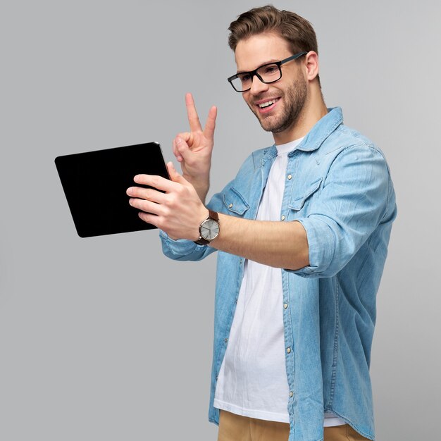 Happy young man wearing jeans shirt standing and using tablet over studio grey wall