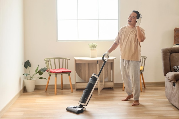 Happy young man wearing wireless headphones cleaning home with vacuum cleaner