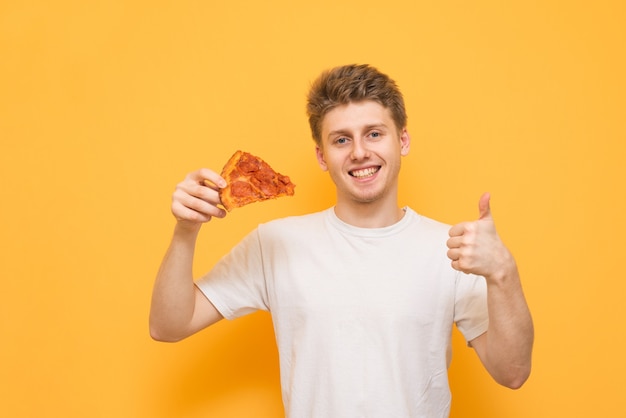 白いTシャツを着た幸せな若い男が彼の手で食欲をそそるピザの一部を保持しています。