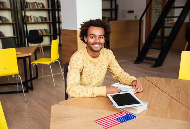 Happy young man with blank screen tablet sitting and reading book in library