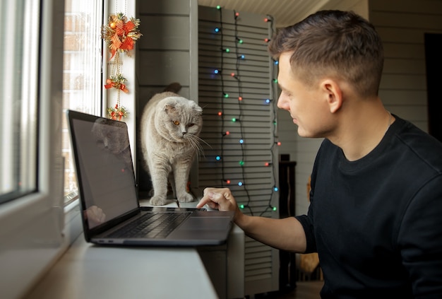 Happy young man with cat at home using zoom to congratulate merry christmas his relatives