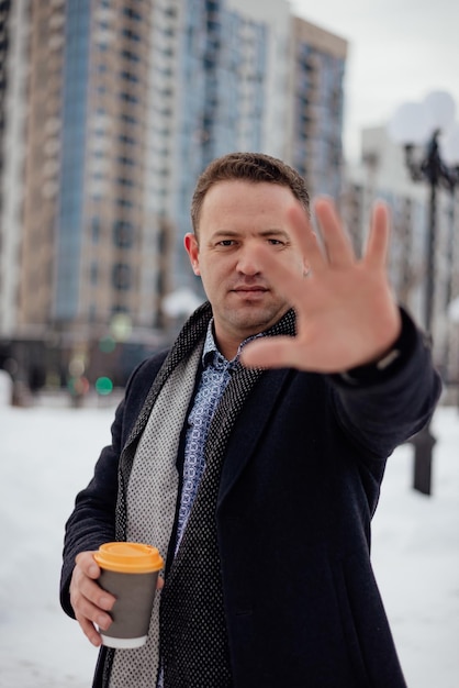 Happy young man with coffee in his hands holding out his hand with an alluring smile portrait looking at the camera vertical photo winter