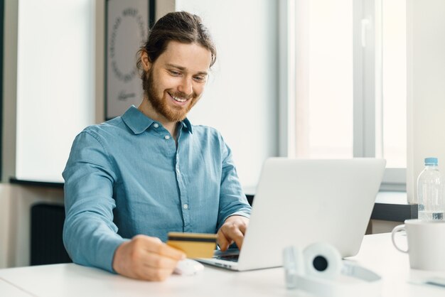 Giovane felice con la carta di credito in mano che fa shopping online sul laptop