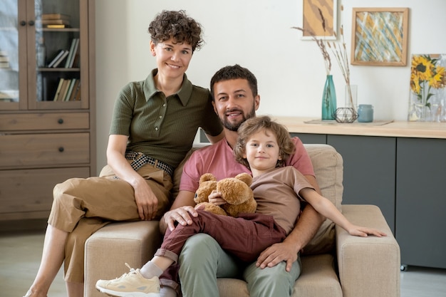 Felice giovane con grazioso piccolo figlio e la sua graziosa moglie seduti in grande poltrona morbida e confortevole al centro del soggiorno di casa