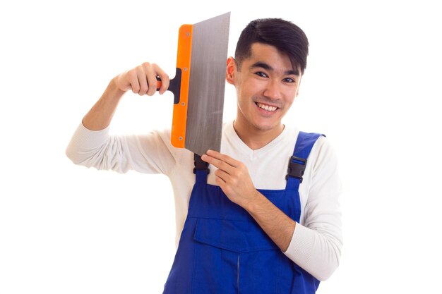 Happy young man with dark hair in white shirt and blue overall holding spatula on white background in studio