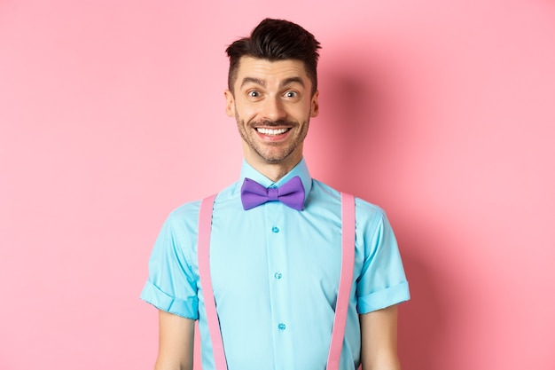 Happy young man with moustache smiling at camera, looking excited and cheerful, standing on pink in bow-tie and shirt.