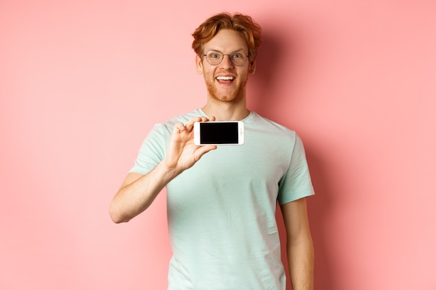 Happy young man with red hair showing smartphone screen holding phone horizontally and smiling amaze...