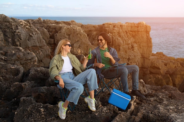 幸せな若い男性と女性がビーチの岩の上でキャンプ椅子でリラックスしています