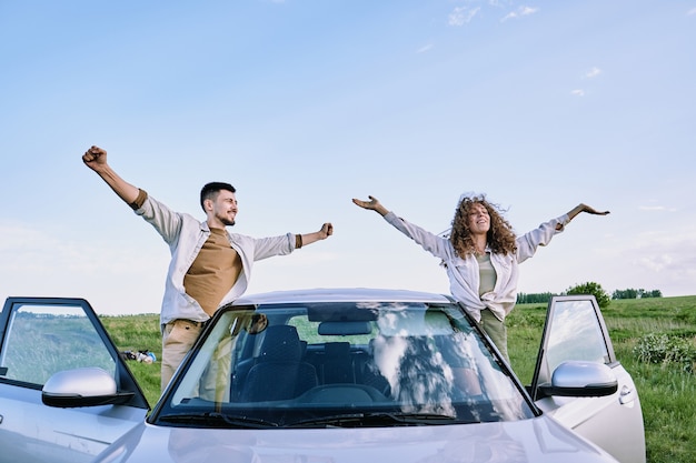 Happy young man and woman with outstretched arms standing by
open doors of their car and enjoying warm summer day against blue
sky