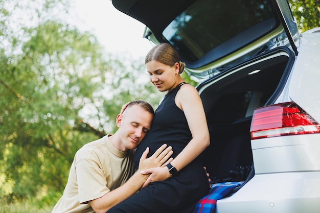 A happy young married couple expecting a child are sitting in the trunk of a car in nature A man and a woman hug a pregnant belly while sitting on a green lawn