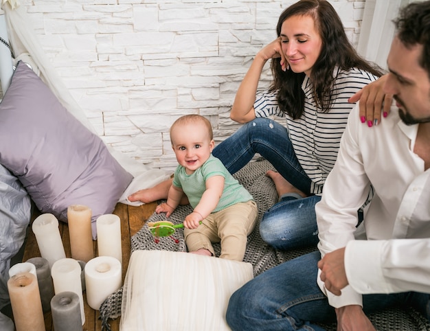 幸せな若い夫婦のポジティブな父と笑顔のお母さんは、彼らの魅力的な幼い息子がガラガラと遊んで床に座っています