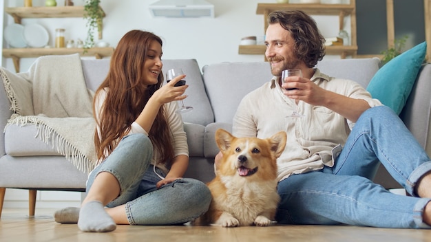 Happy young married couple sitting on the floor near a couch and clinking glasses with wine