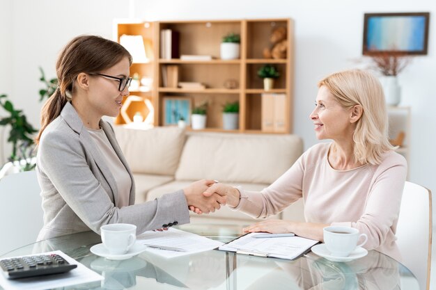 取引後にテーブルの上で握手しながらお互いを見て幸せな若いと成熟したエレガントな女性