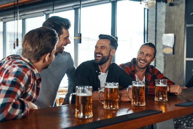 Happy young men in casual clothing drinking beer and laughing while sitting in the pub