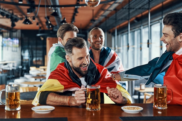 Foto giovani uomini felici coperti di bandiere internazionali che bevono birra e si uniscono mentre sono seduti al pub