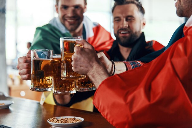 パブでスポーツの試合を見ながらビールで乾杯する国際的な旗で覆われた幸せな若い男性