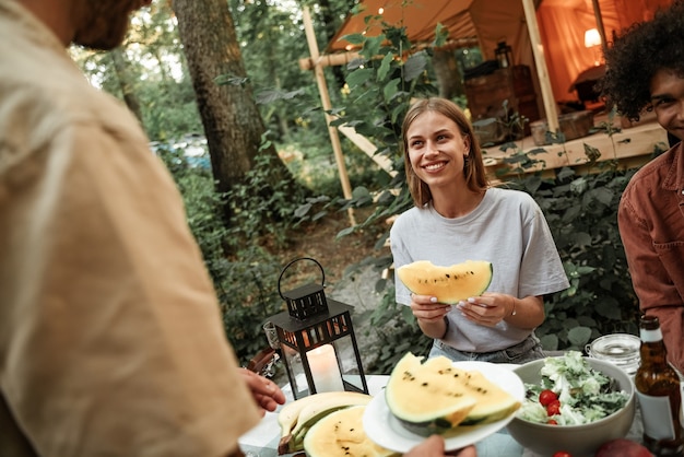 Foto felice giovane donna millenaria con i capelli biondi che sorride con i denti bianchi agli amici che tengono una fetta di cocomero. estate piena di vitamina, concetto di frutta esotica. dieta sana, cibo glamping, zucchero non raffinato