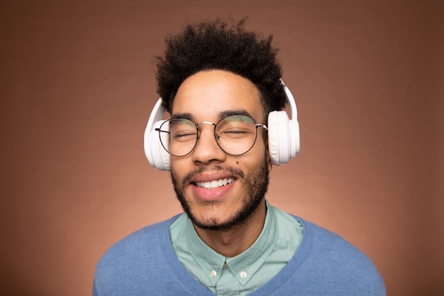 ヘッドフォンで音楽を楽しみながら目を閉じたまま眼鏡とスマートカジュアルウェアで幸せな若い混血の実業家