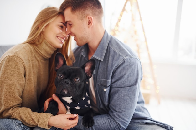 Photo happy young modern couple sitting on the sofa at home with christmas tree with their cute dog.