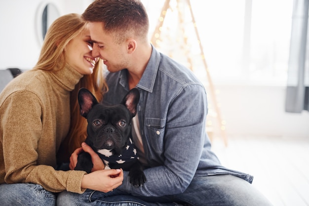 Happy young modern couple sitting on the sofa at home with christmas tree with their cute dog.