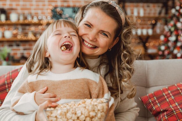 행복한 젊은 엄마와 아기 소녀는 소파에 앉아 팝콘을 먹으며 웃고 있습니다. TV를 보고 팝콘을 먹는 가족.