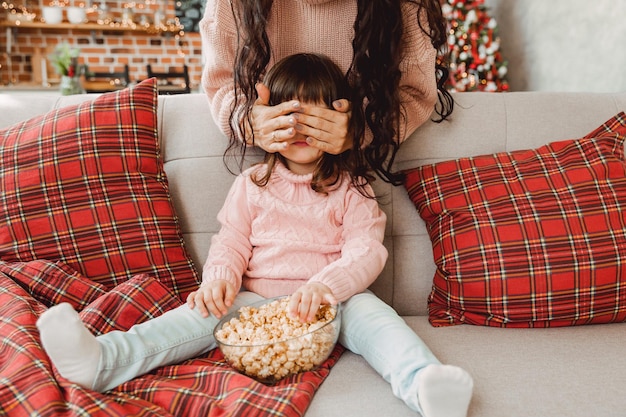 幸せな若いお母さんと女の赤ちゃんは、ソファに座ってポップコーンを食べながら笑います。家族はテレビを見たり、ポップコーンを食べたりします。