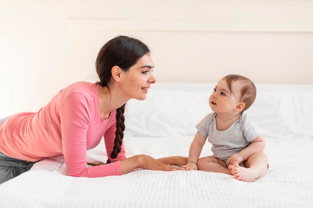 Happy young mom resting with baby girl on bed adorable child\
sitting and looking at mom spending time together