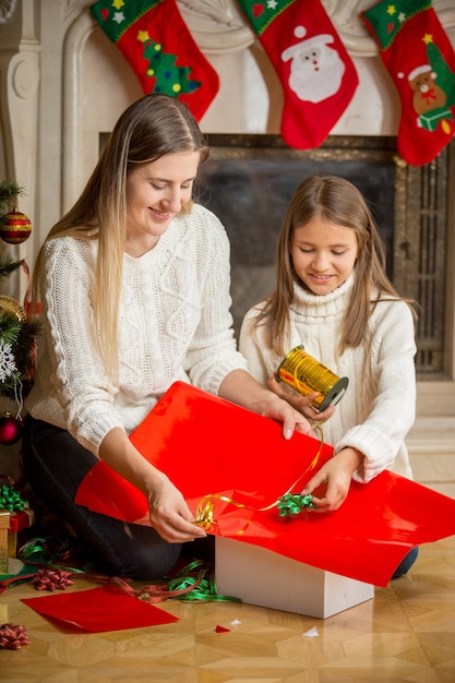 Felice giovane madre e figlia carina che confezionano regali di natale in carta rossa e li legano con un nastro dorato