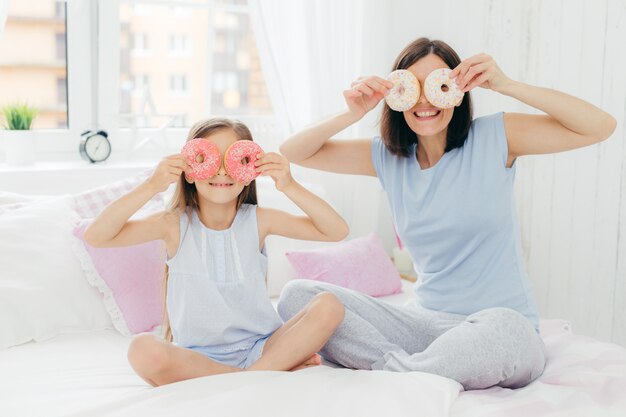 Happy young mother and daughter have fun together, hold tasty sweet doughnuts near eyes