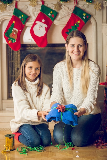 Happy young mother and daughter sitting on floor at fireplace and packing sweater for Christmas