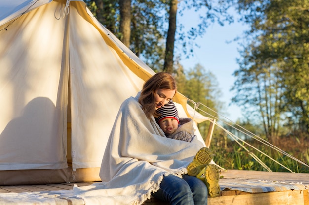 Happy young mother embracing her toddler girl with a blanket while sitting near canvas tent