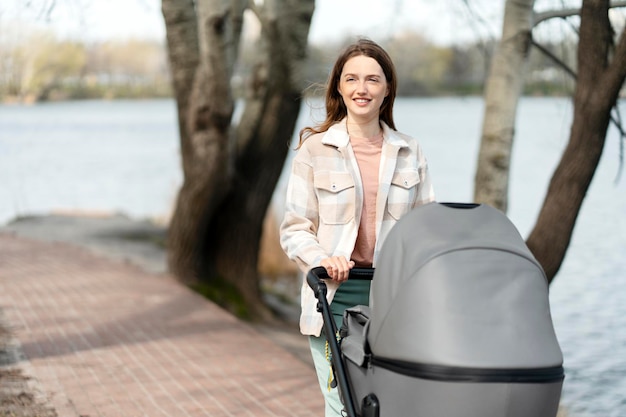 Happy young mother pushing stroller with little child walking in nature outdoors near lake