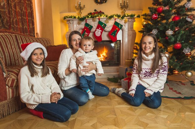 Happy young mother sitting with children on the floor at fireplace. Decorated Christmas tree on background.