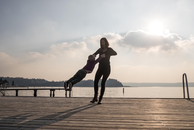 Happy young mother standing on wooden pier holding her toddler son by his hands spinning him around with ocean in background.