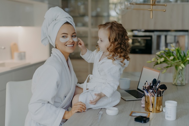 Happy young mother teacher her little daughter to do makeup.