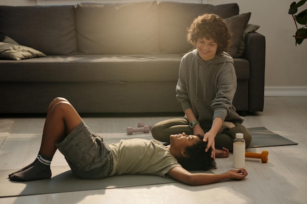 Happy young mother touching hair of her son relaxing on mat on the floor