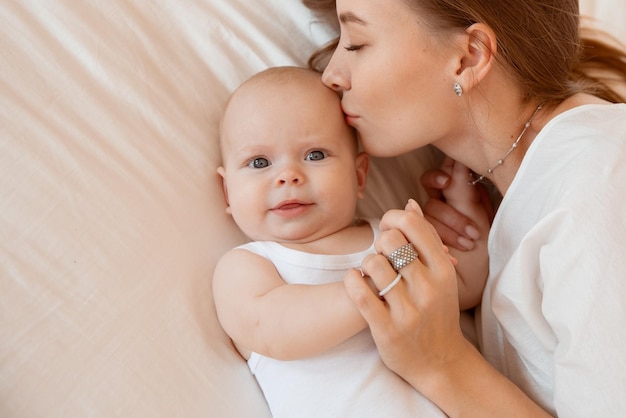 하얀 방에 아기를 안고 있는 행복한 젊은 엄마는 침실에 있는 집에서 아기에게 키스를 하고 있다