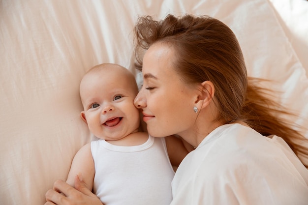 하얀 방에 아기를 안고 있는 행복한 젊은 엄마는 침실에 있는 집에서 아기에게 키스를 하고 있다