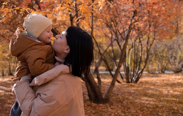 A happy, young mother with a child in an autumn park. Mom holds hugs and kisses her son on the street. Autumn season, the concept of parenting and children. Place to copy