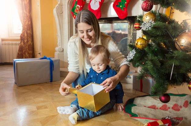 아기 아들이 크리스마스 트리에 앉아 선물 상자 안을 들여다보는 행복한 젊은 어머니