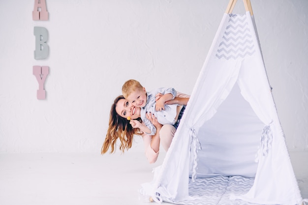 Happy young mother with her smiling son looks out of a child's wigwam in the light room.