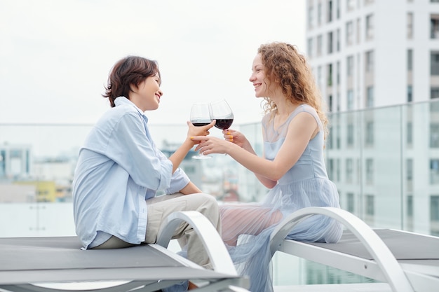 Happy young multi-ethnic lesbian couple drinking wine when having romantic date on rooftop