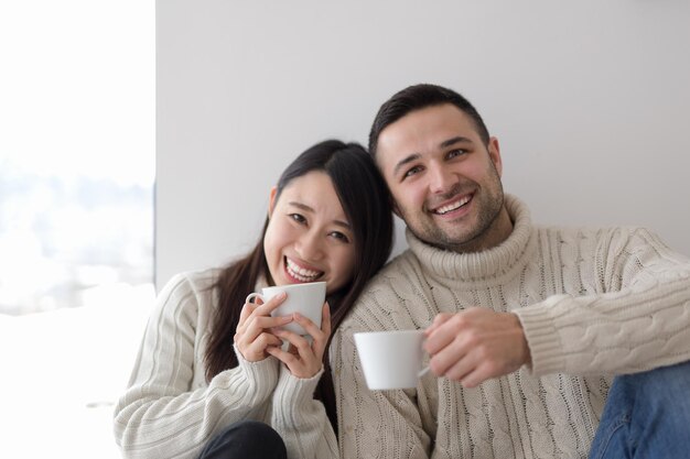 추운 겨울날 집에서 창가에서 모닝 커피를 즐기는 행복한 젊은 다민족 커플