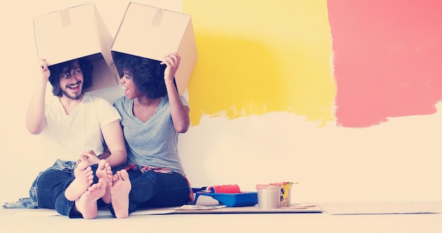 Happy young multiethnic couple relaxing and playing with cardboard boxes after painting a room in their new house on the floor