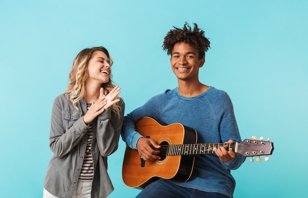 Happy young multiethninc couple together, playing a guitar isolated over blue wall