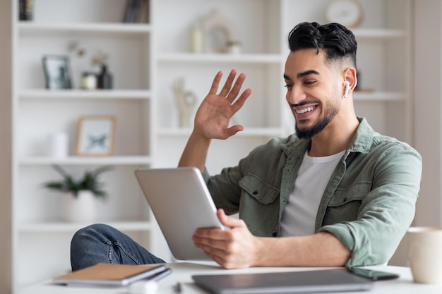 Happy young muslim guy with beard in wireless headphones looks at tablet and waves his hand