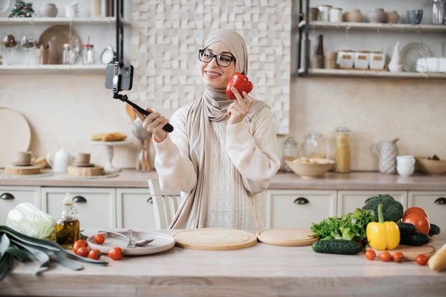 Foto felice giovane donna musulmana che utilizza lo smartphone in bastone selfie in piedi in cucina al mattino