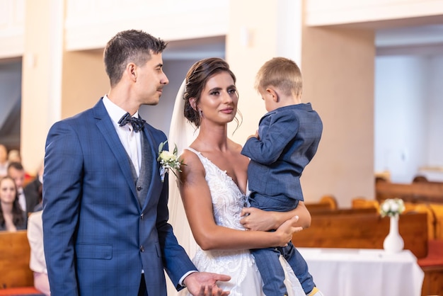 Happy young newlyweds stand in the church with the bride holding a young son in her arms